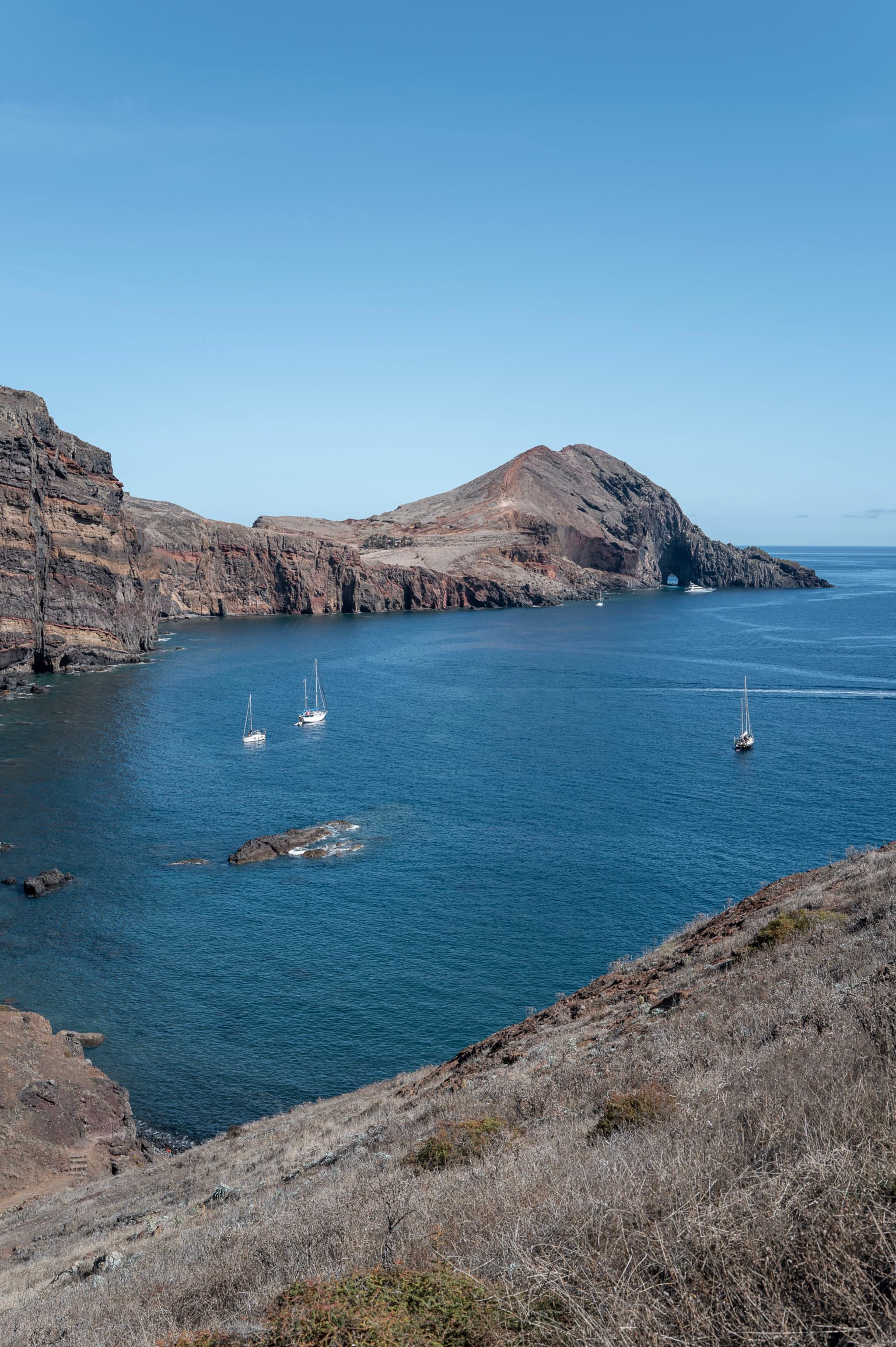 A rocky coastline with several sailboats in a calm blue bay under a clear sky