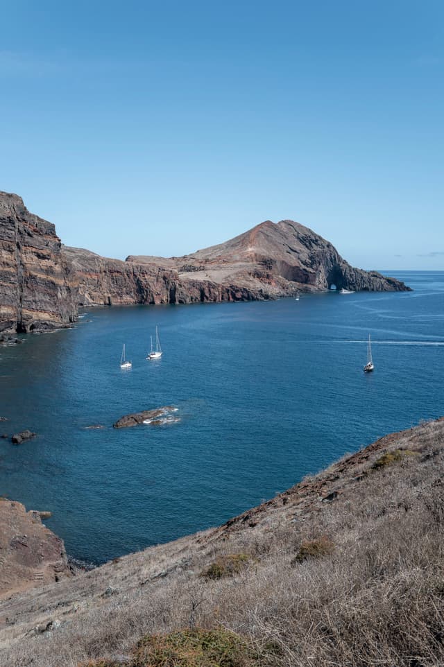 A rocky coastline with several sailboats in a calm blue bay under a clear sky