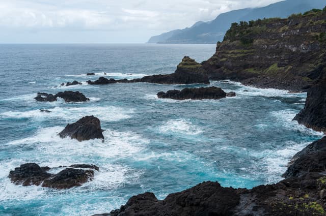Rugged cliffs along a rocky coastline with waves crashing against them, under an overcast sky