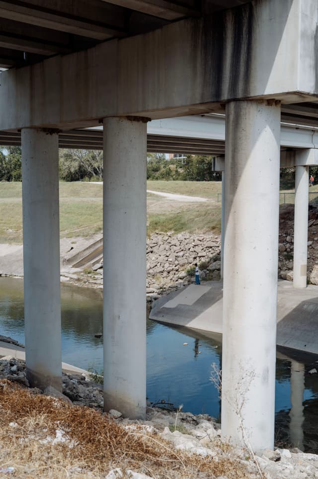 A concrete bridge with large pillars spans over a small body of water, with grassy areas and a person standing near the water's edge