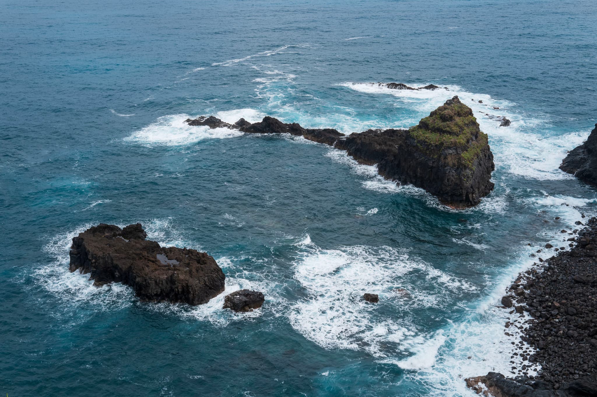 Rocky outcrops in a turbulent ocean with waves breaking against them, under a cloudy sky