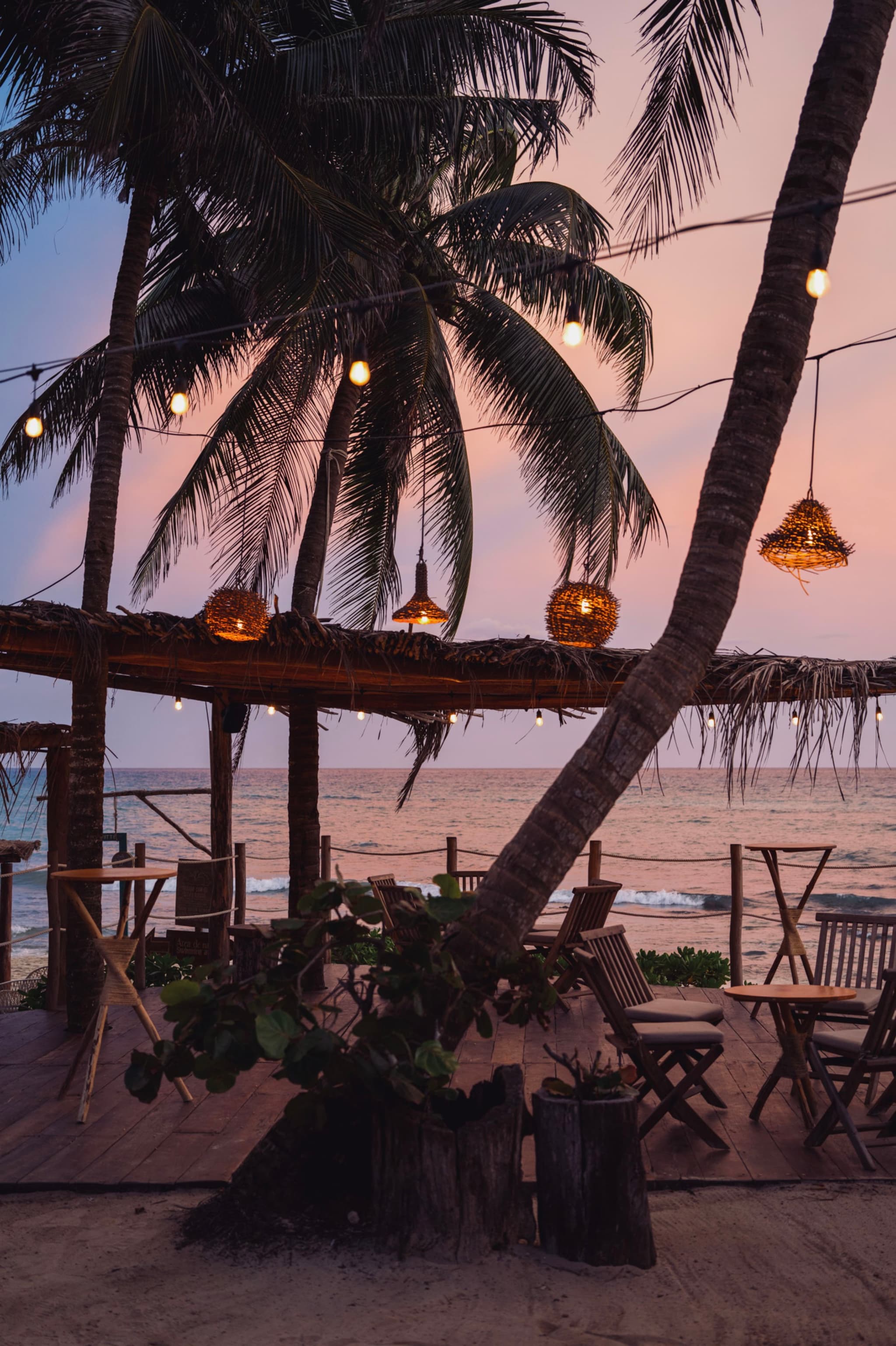 A beachside setting with palm trees, string lights, and wooden tables and chairs under a thatched roof, overlooking the ocean at sunset