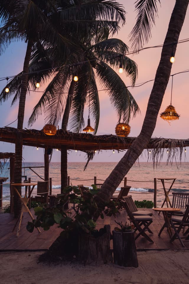 A beachside setting with palm trees, string lights, and wooden tables and chairs under a thatched roof, overlooking the ocean at sunset