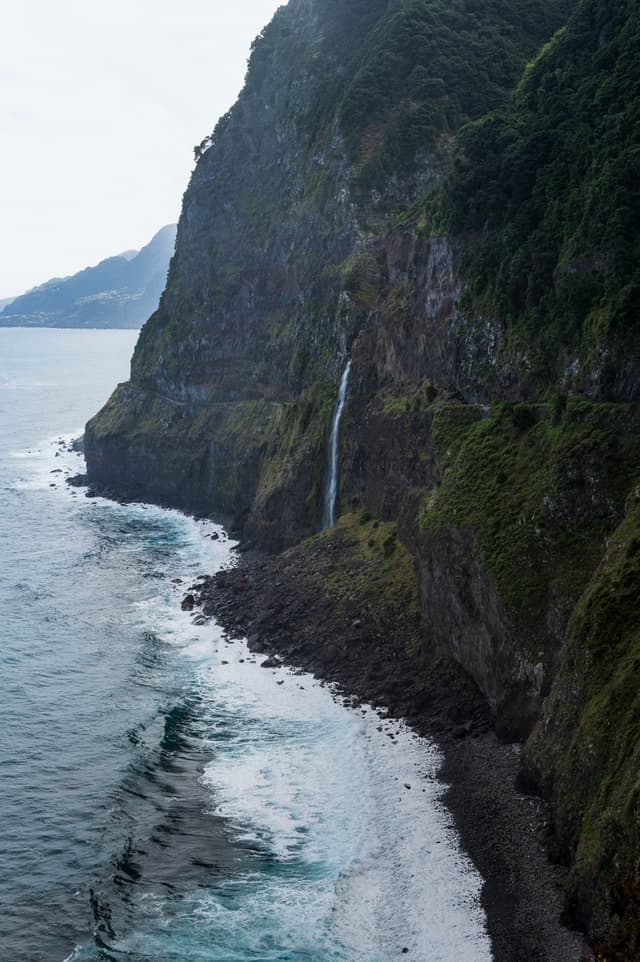 A narrow waterfall cascades down a steep, forested cliff into the ocean, with waves lapping at a rocky shoreline