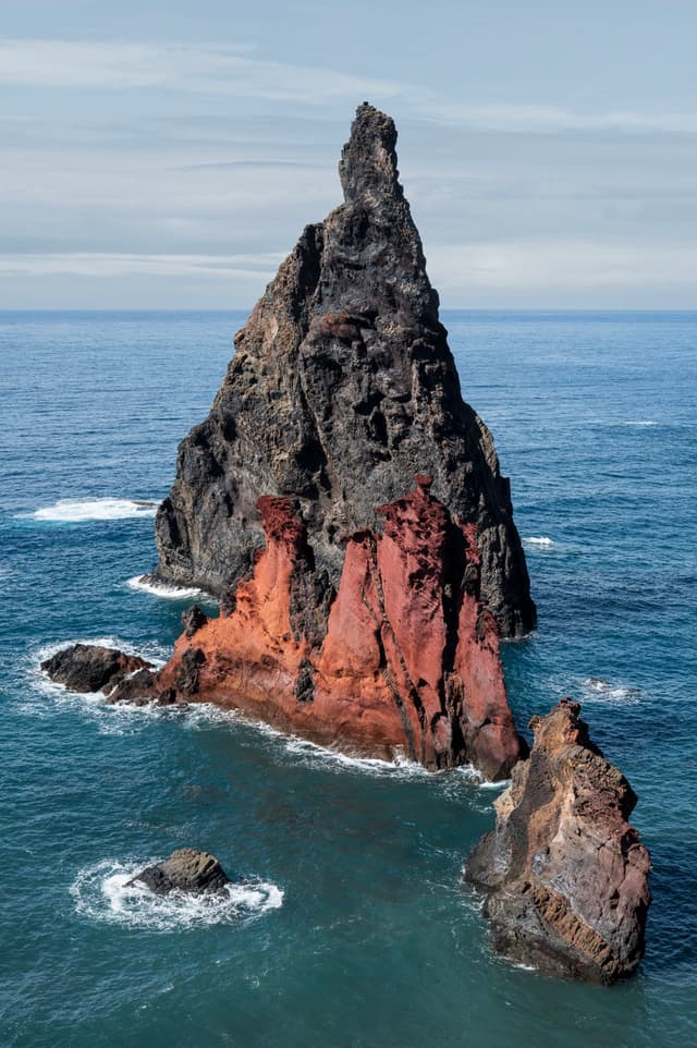 Tall, jagged rock formation rising from the ocean with vibrant red and dark tones, surrounded by blue water and a smaller rock in the foreground