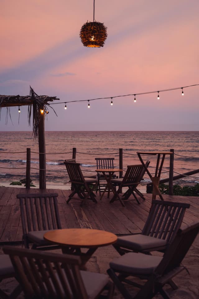 A seaside deck with wooden chairs and tables, string lights, and a hanging lantern, set against a pink and purple sunset sky