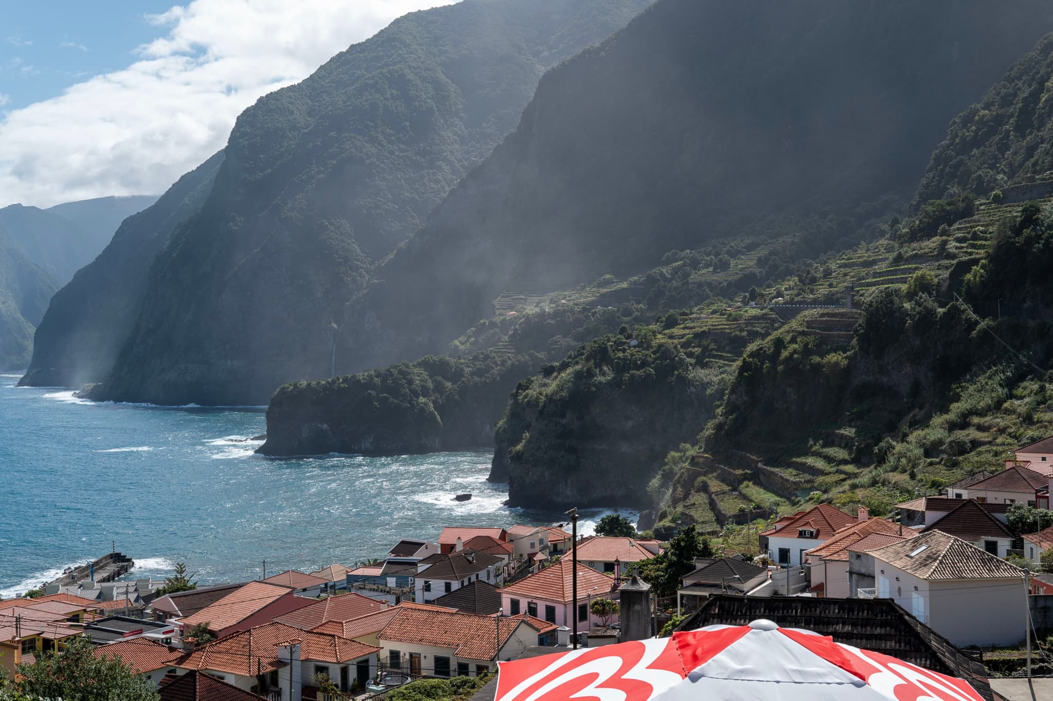 Coastal village with terracotta roofs, misty green mountains, and rugged coastline under a partly cloudy sky
