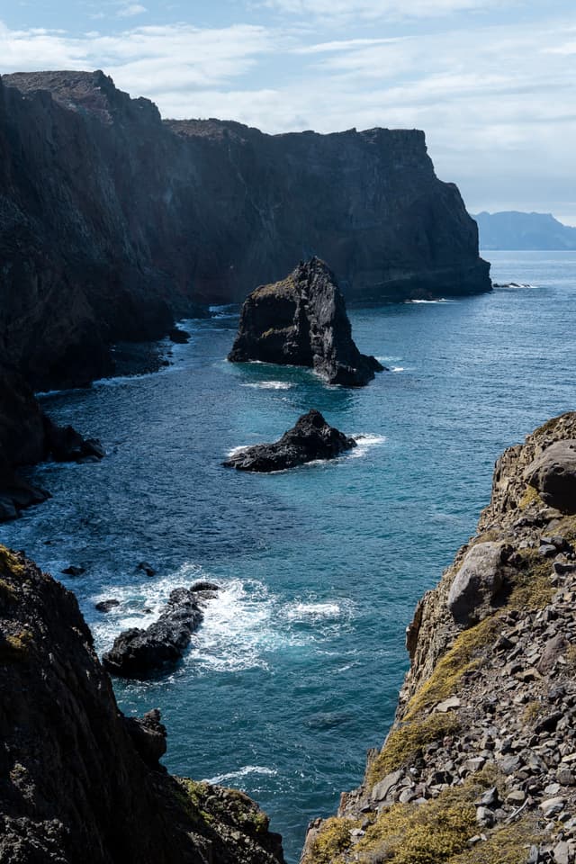 Rugged cliffs surrounding deep blue ocean waters, with isolated rock formations jutting out and partially cloudy sky overhead
