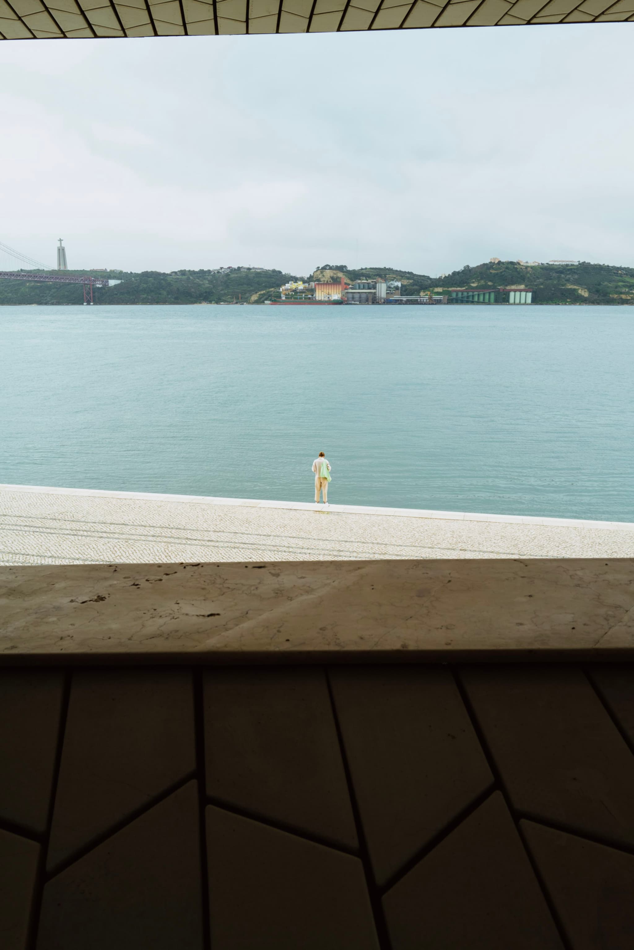 A person stands on a wide, light-colored platform overlooking a calm body of water, with a distant shoreline and buildings visible under a cloudy sky. The scene is framed by geometric patterns in the foreground
