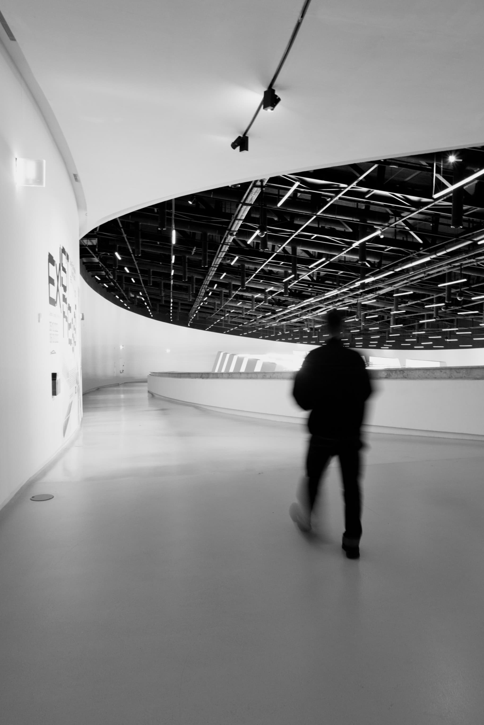 A person walking through a modern, curved hallway with a sleek, minimalist design and overhead lighting. The image is in black and white, emphasizing the architectural lines and shadows