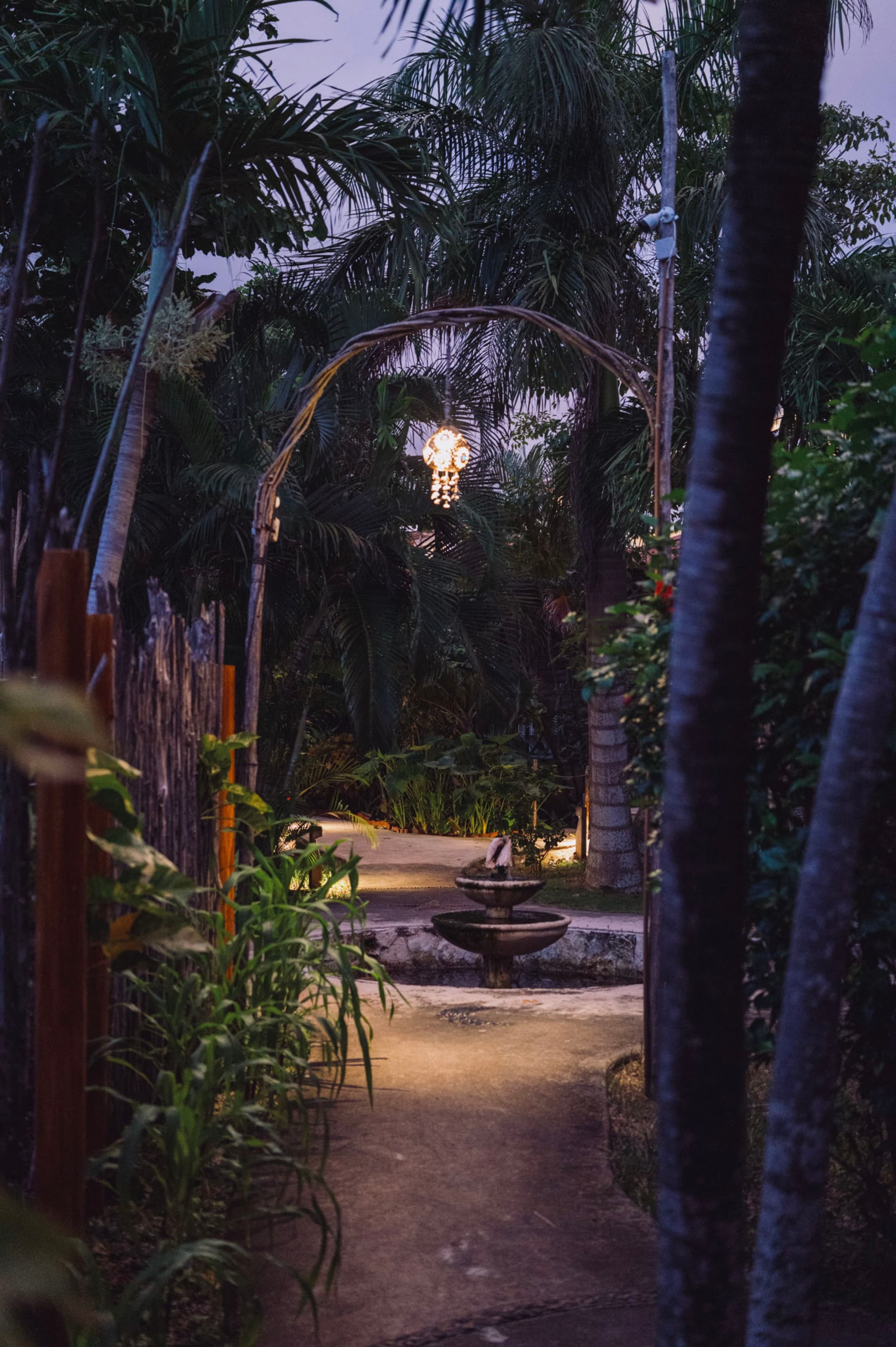 A dimly lit garden path with tropical plants, a wooden archway, and a small fountain, illuminated by a hanging light