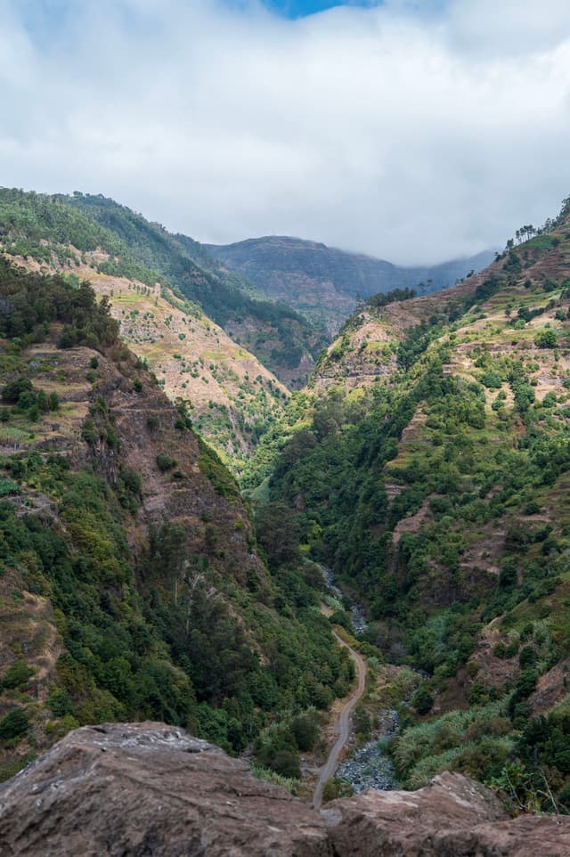 Lush green valley with rugged cliffs, winding path, and cloudy sky in a mountainous landscape