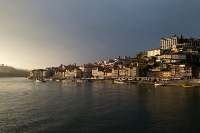 A coastal cityscape at sunset with buildings along the waterfront and a calm body of water in the foreground