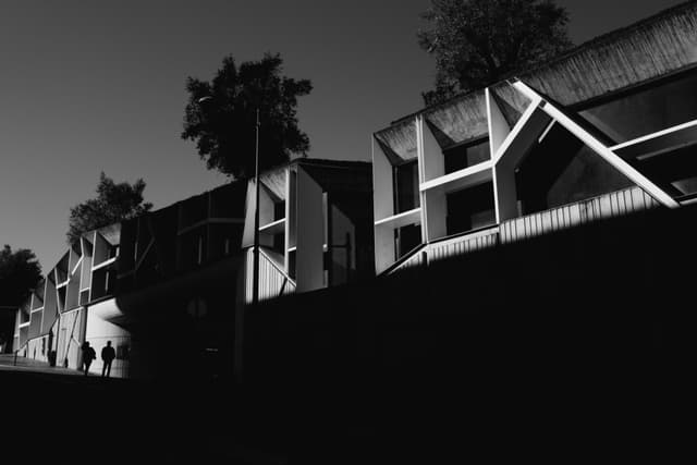 A black and white scene featuring modern architectural buildings with geometric designs, casting sharp shadows. Silhouetted trees and people are visible against a clear sky