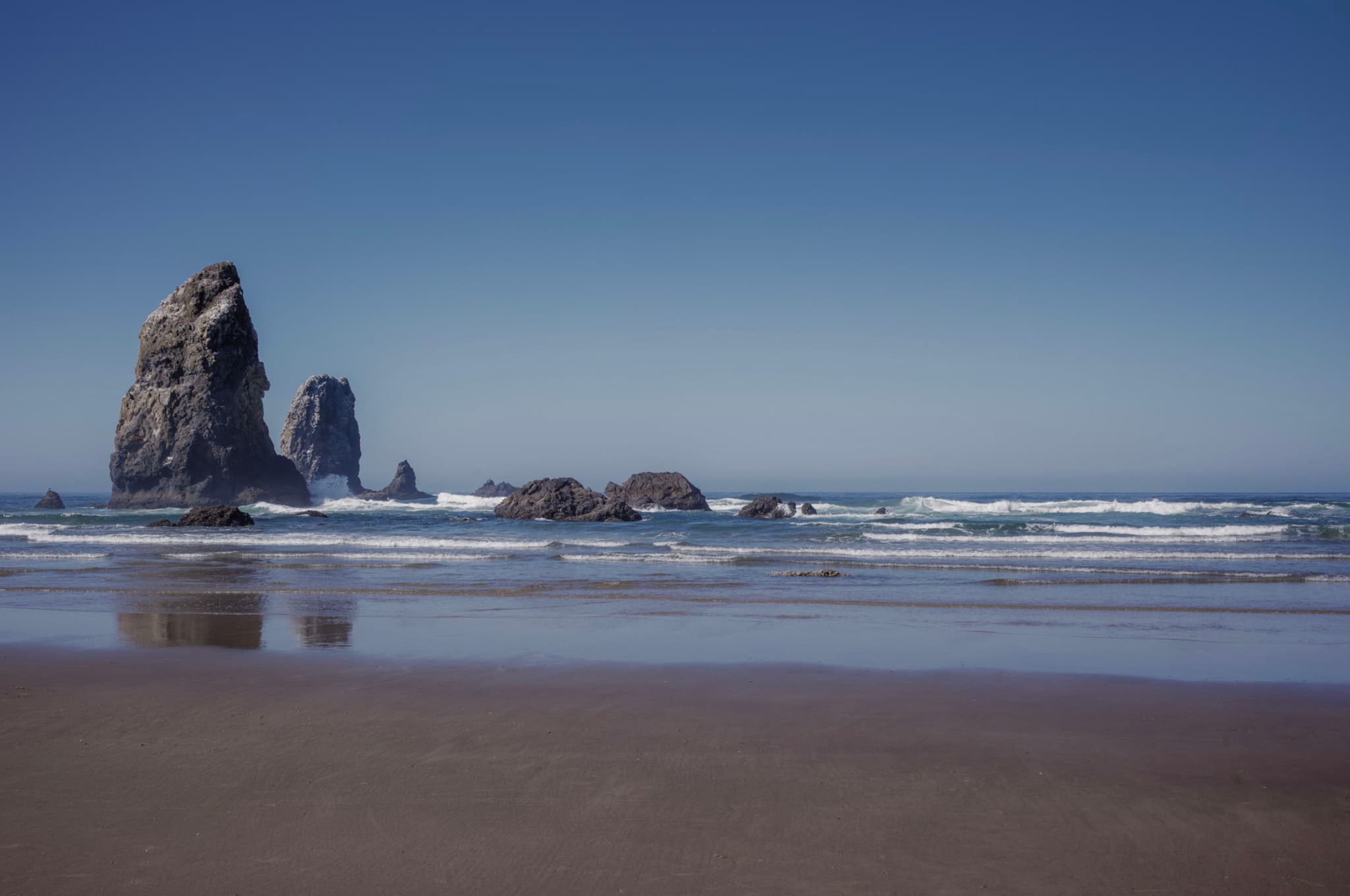 A serene beach scene with large rock formations in the ocean, gentle waves, and a clear blue sky