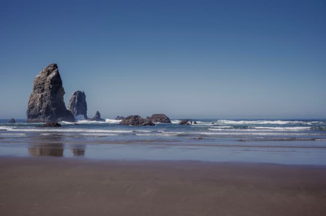 A serene beach scene with large rock formations in the ocean, gentle waves, and a clear blue sky
