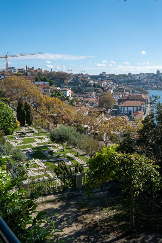 A scenic view of a hillside garden with geometric patterns, overlooking a cityscape and a river under a clear blue sky
