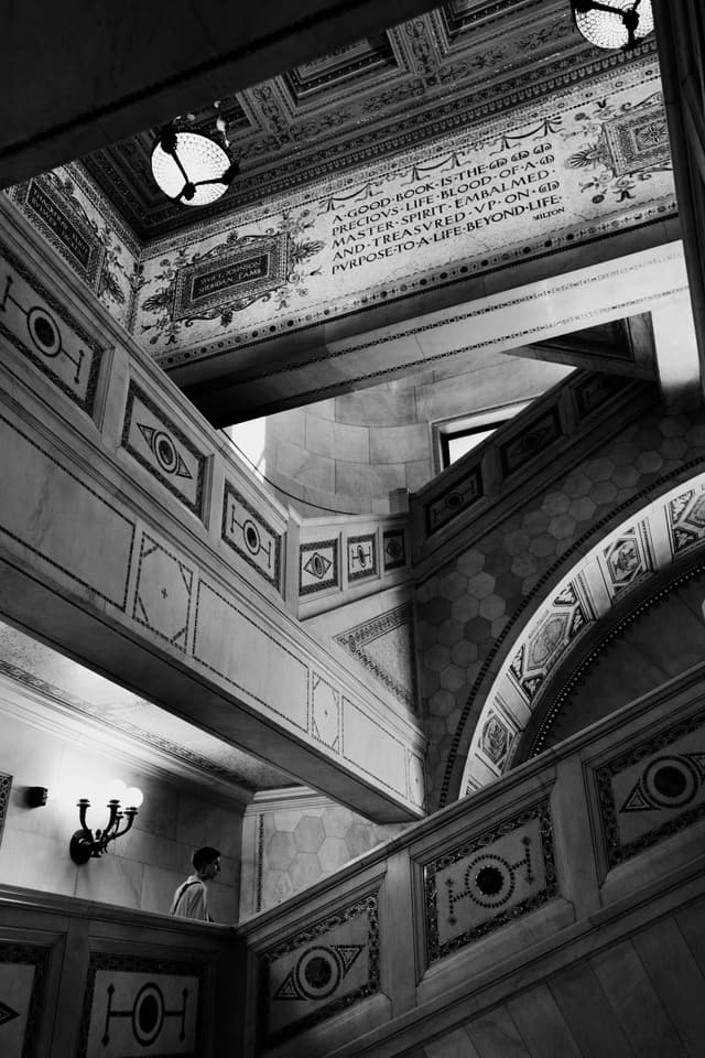 A black and white photograph of an ornate, multi-level staircase with decorative patterns and inscriptions on the walls and ceiling. A person is visible on one of the staircases