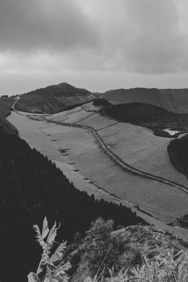 A black and white landscape featuring a winding road through hilly terrain, with clouds overhead and vegetation in the foreground