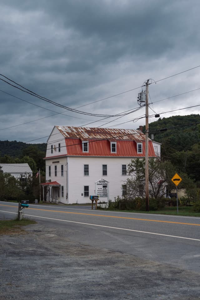 A white building with a red roof near a road, under a cloudy sky with visible power lines
