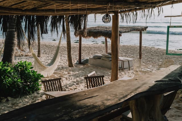 A rustic beach setting with wooden furniture, a hammock, and a thatched roof, overlooking the ocean