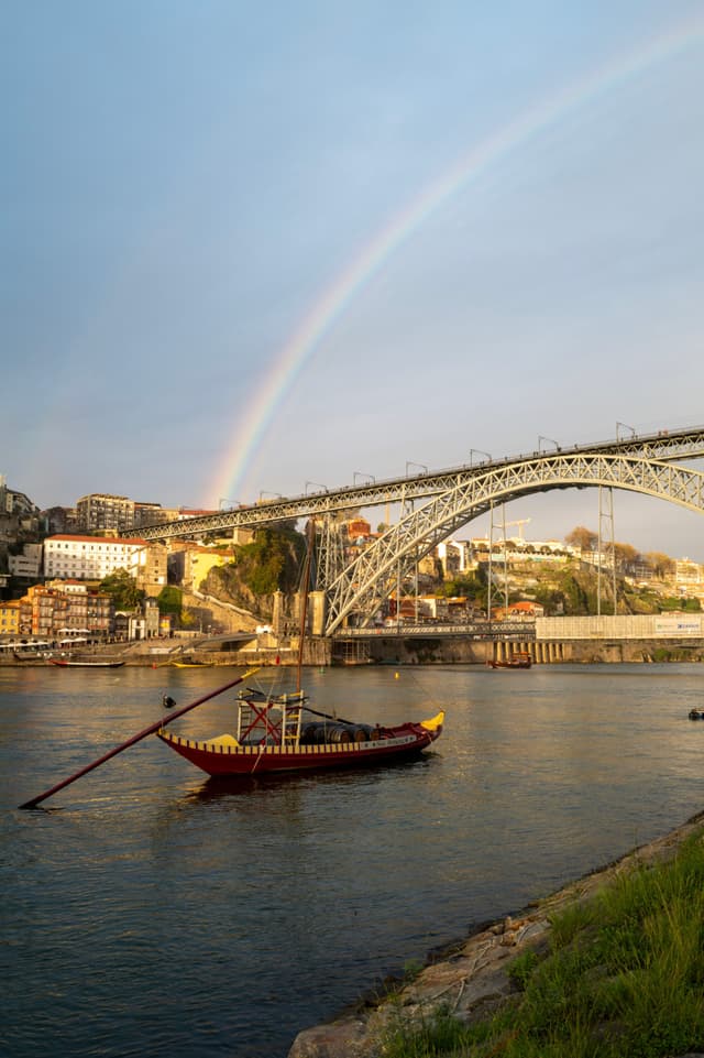A traditional boat floats on a river beneath a large arched bridge, with a rainbow arching over a cityscape in the background