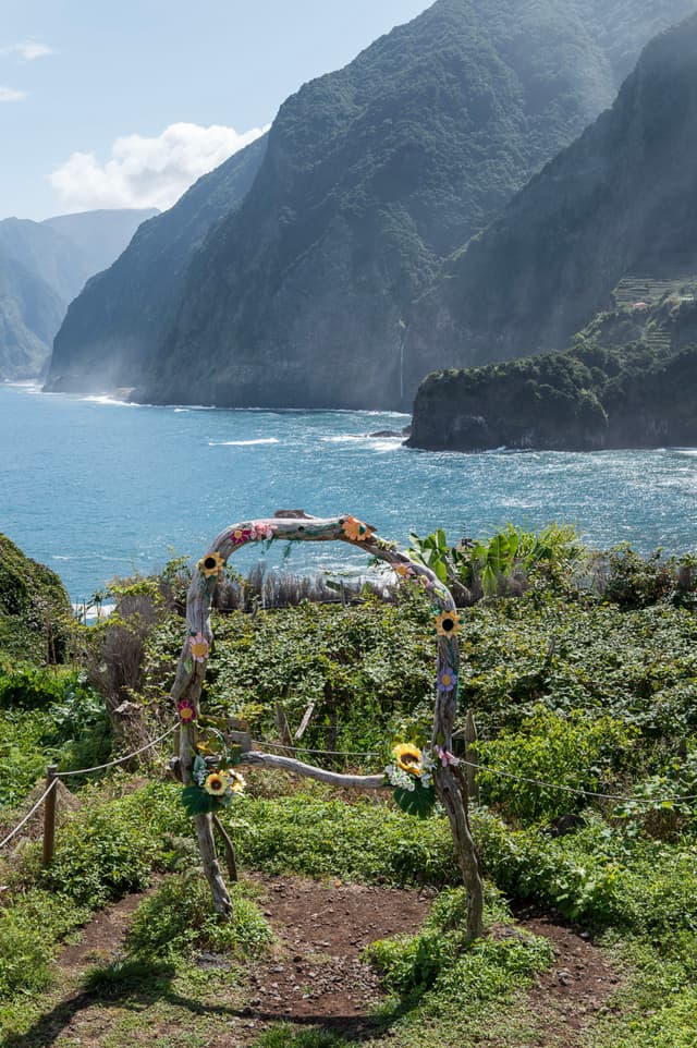 A scenic cliffside with a custom floral arch overlooking a blue ocean and distant mountains under a clear sky