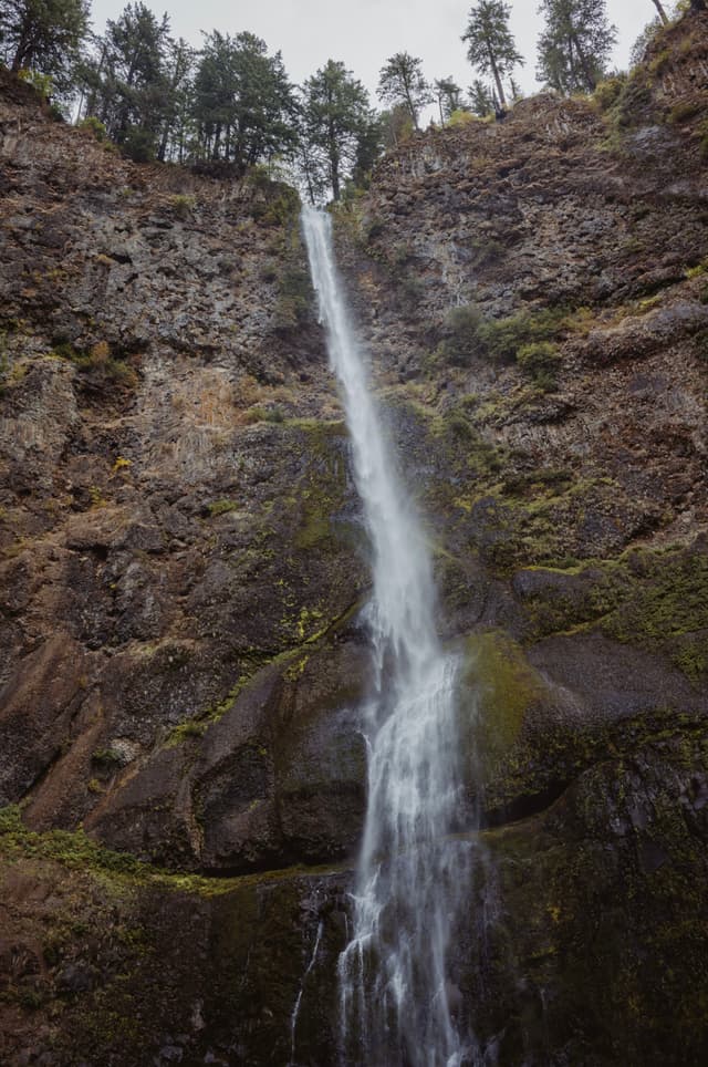 A tall, narrow waterfall cascades down a rocky cliff surrounded by trees at the top