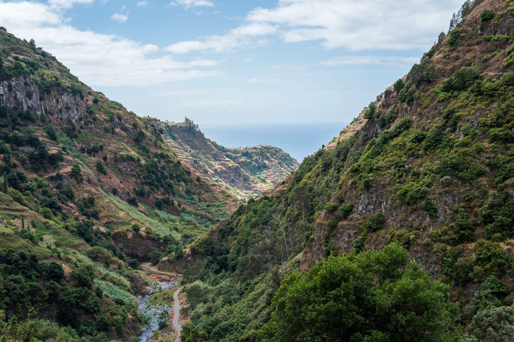 A lush green valley flanked by steep hills, opening to a view of the sea under a partly cloudy sky