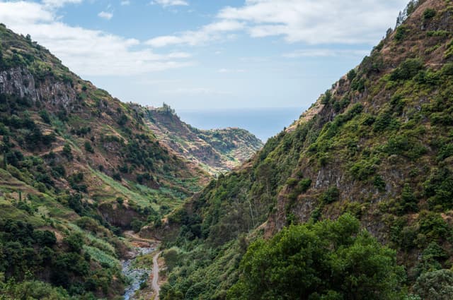 A lush green valley flanked by steep hills, opening to a view of the sea under a partly cloudy sky