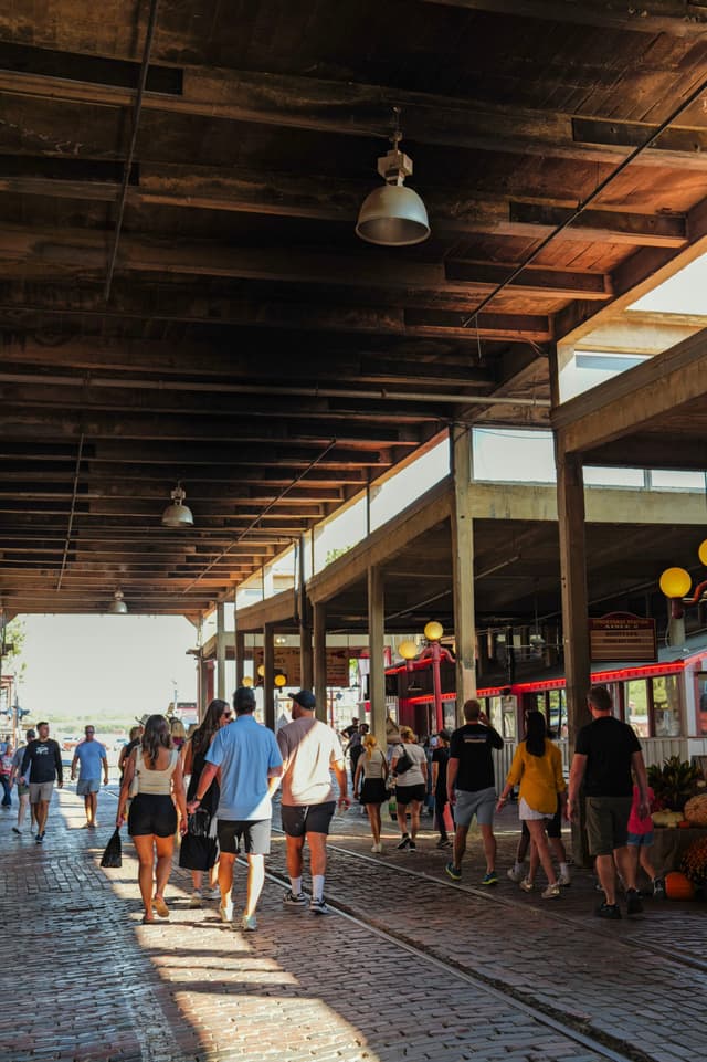 A group of people walking under a covered walkway with a wooden ceiling and exposed beams, surrounded by shops and bright lighting