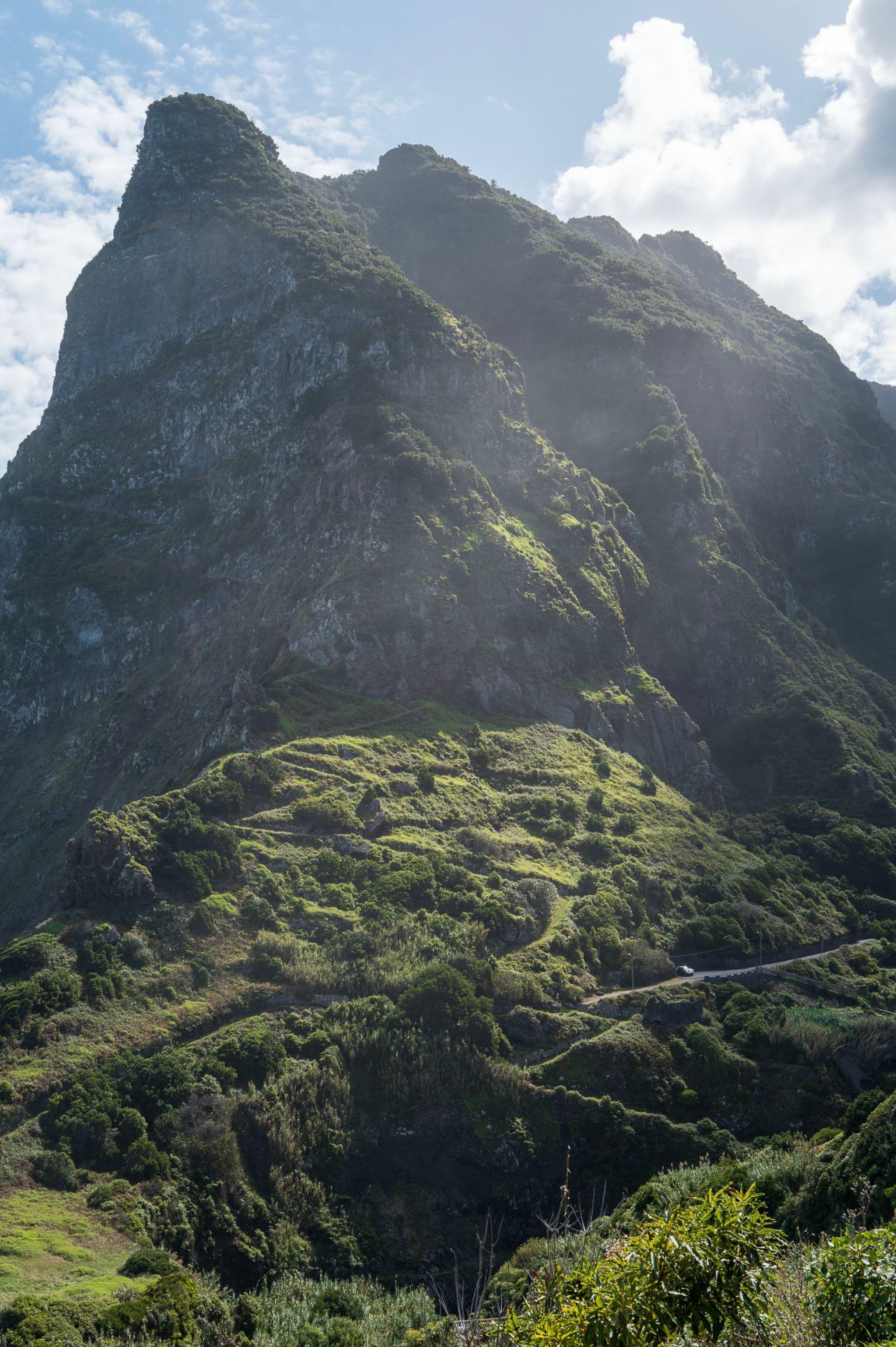 Steep, lush green mountain slope with winding roads under a bright blue sky and scattered clouds