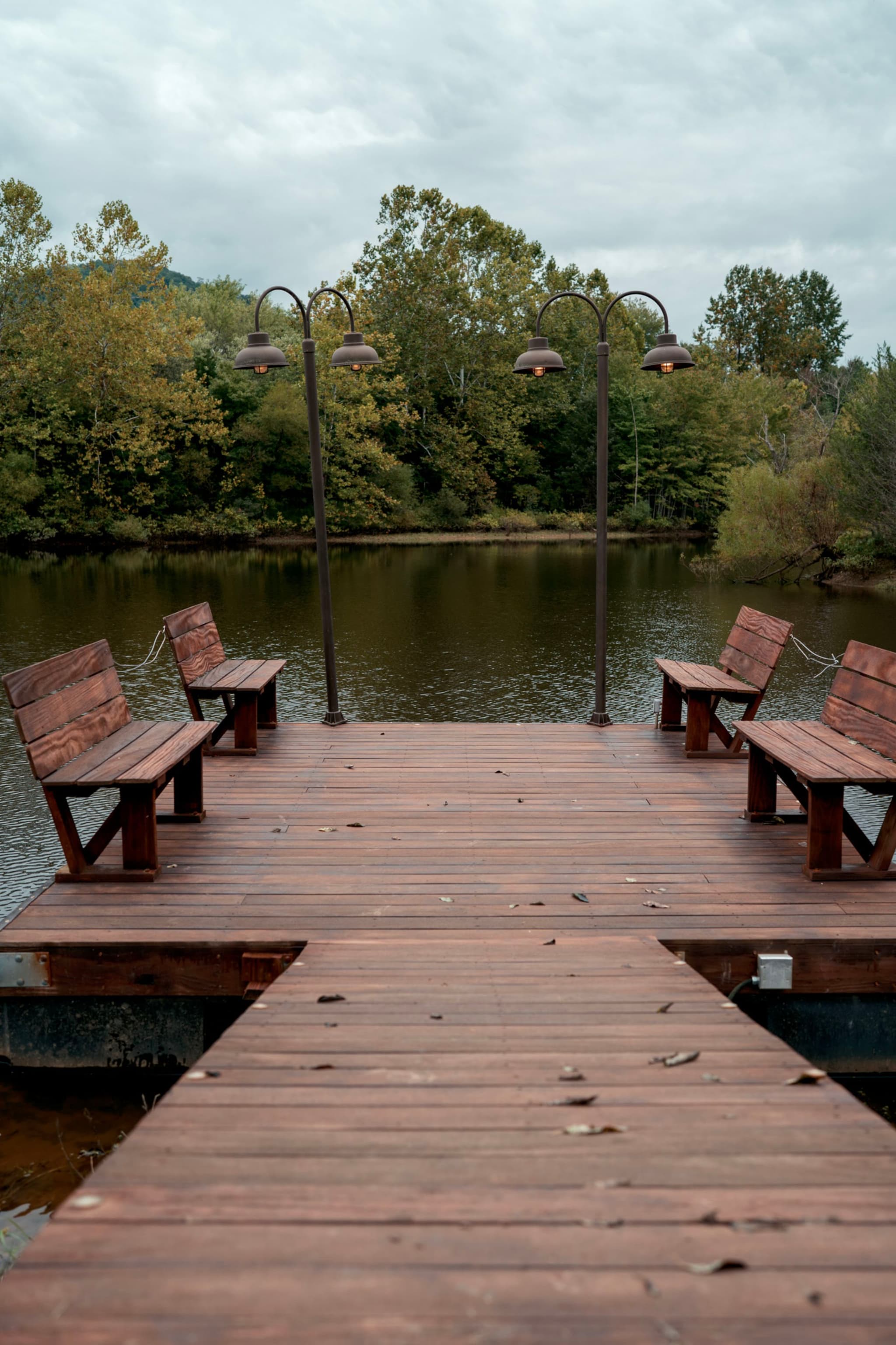 A wooden dock extends over a calm lake, flanked by benches and lit by vintage-style lamps, with a backdrop of trees under a cloudy sky