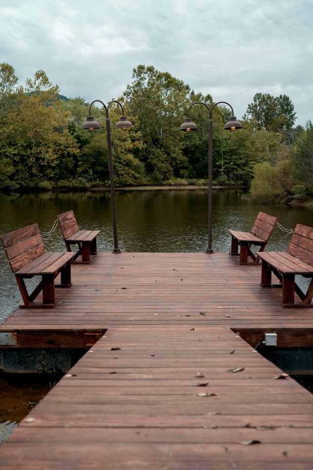 A wooden dock extends over a calm lake, flanked by benches and lit by vintage-style lamps, with a backdrop of trees under a cloudy sky