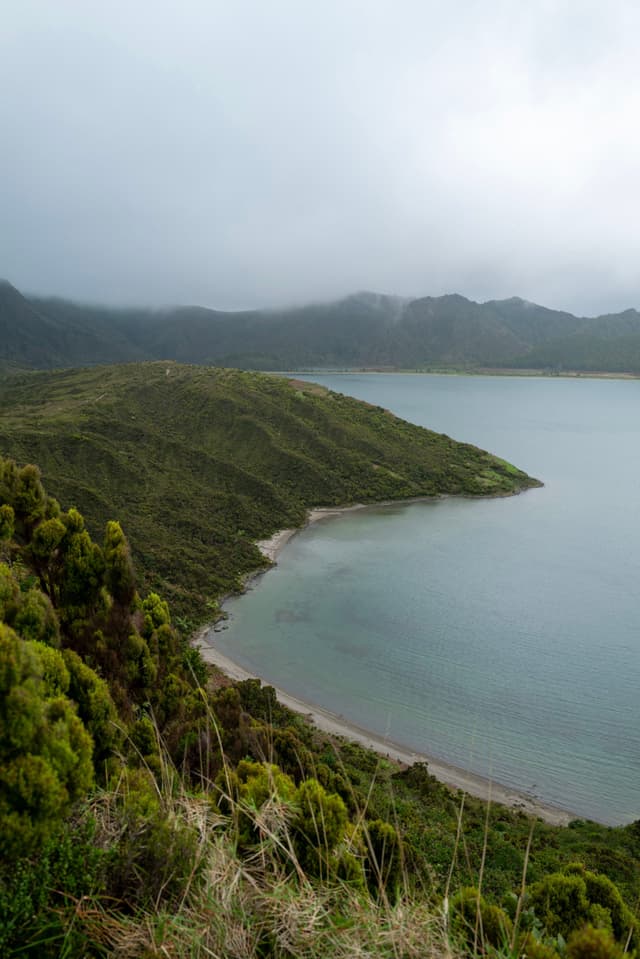 A serene coastal landscape with lush green hills, a calm body of water, and a cloudy sky