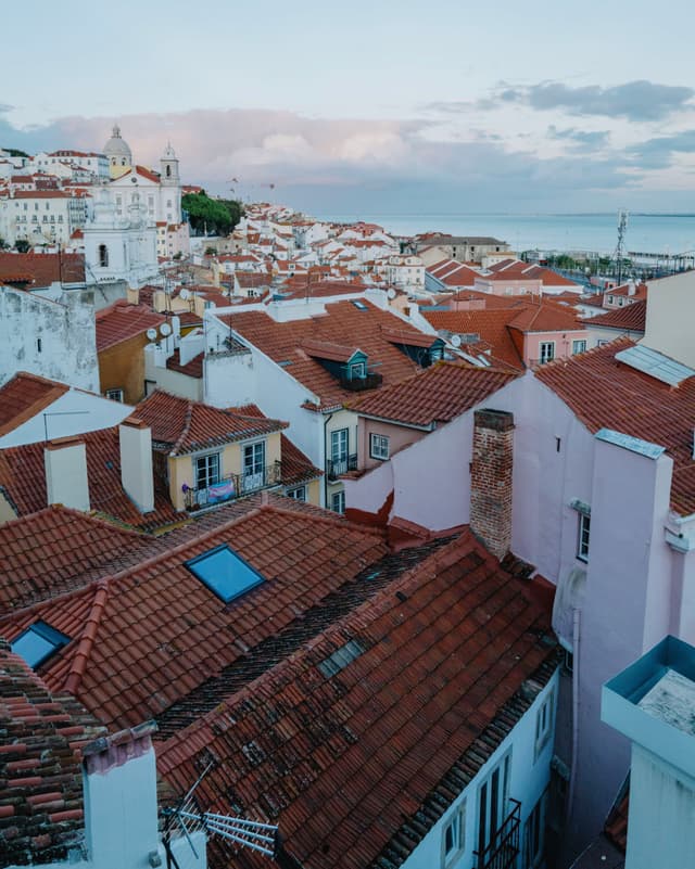 A scenic view of a coastal town with red-tiled rooftops, pastel-colored buildings, and a distant ocean horizon under a cloudy sky