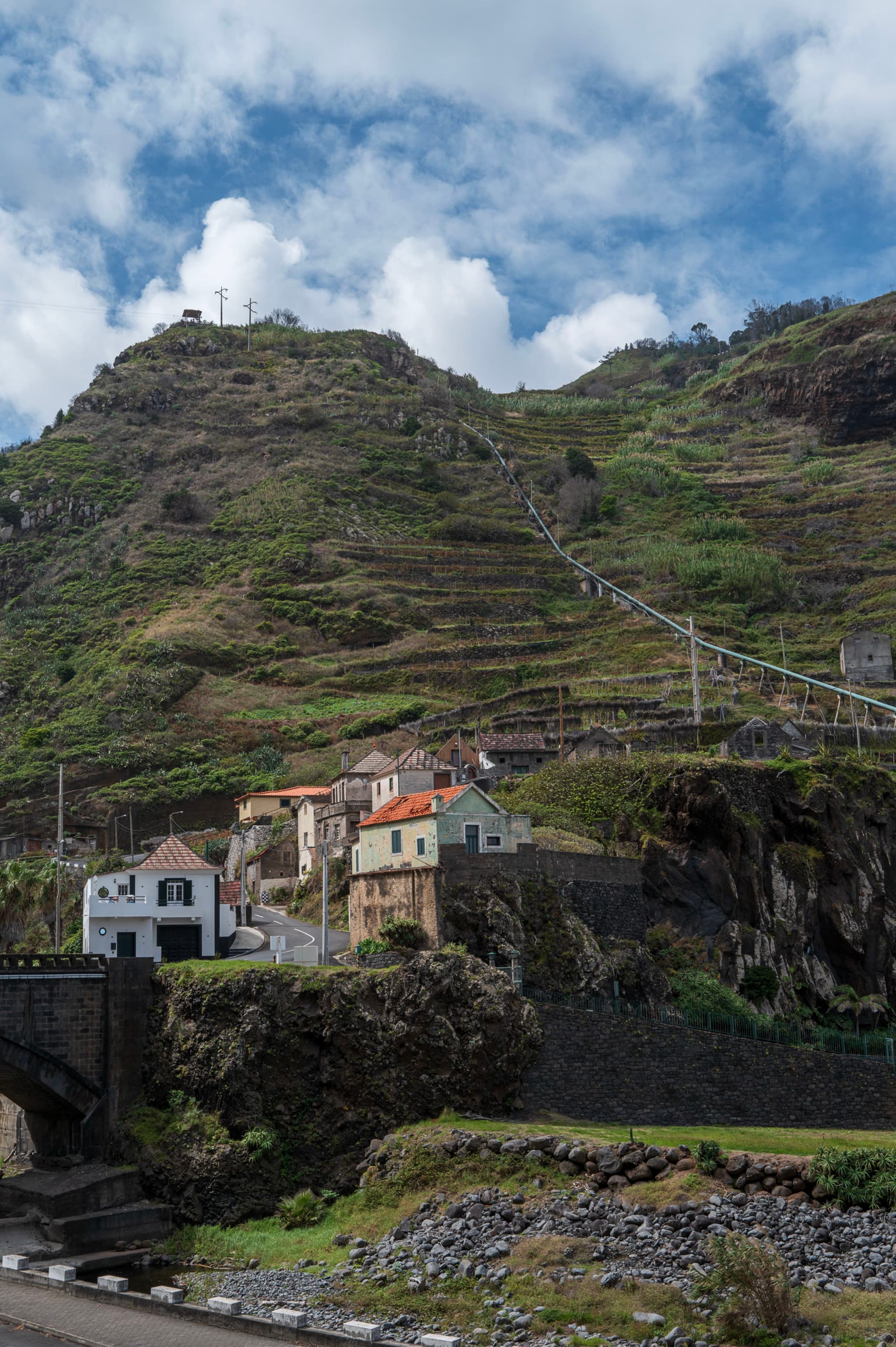 Cluster of houses built on a terraced, green hillside under a partly cloudy sky
